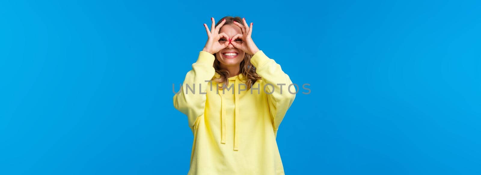 Dreamy and silly girl looking through fingers with happy playful expression, make okay gestures form glasses with hands and smiling carefree, look far away, searching something, blue background.