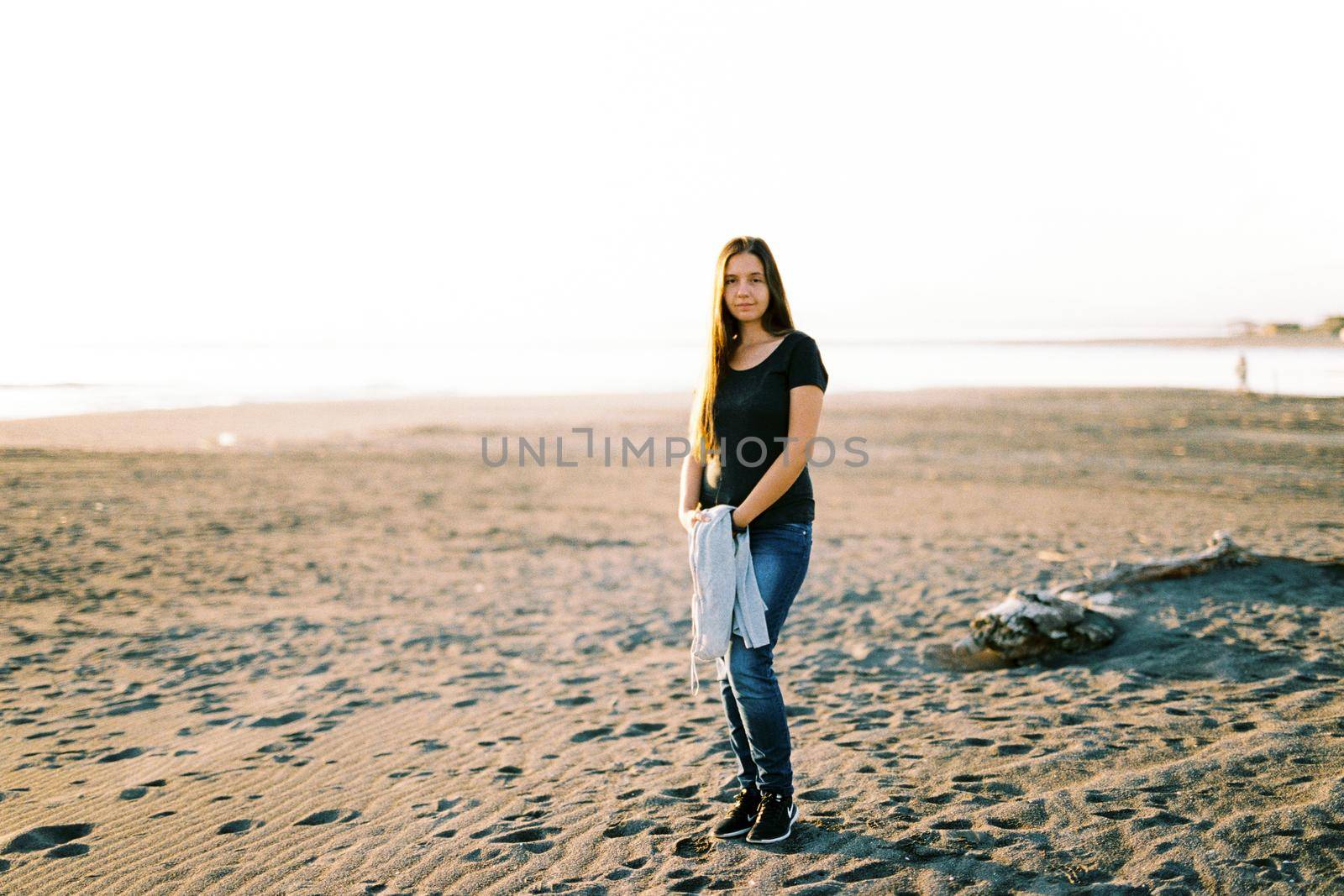 Girl in jeans with a sweater in her hands stands on a sandy beach by the sea. High quality photo