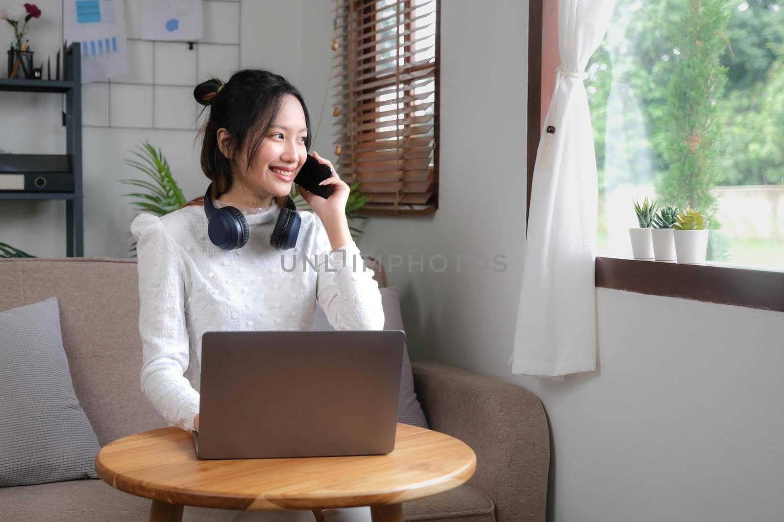 Charming young Asian female relaxes in the living room enjoying talking with her coworker on her phone while working from home. by wichayada