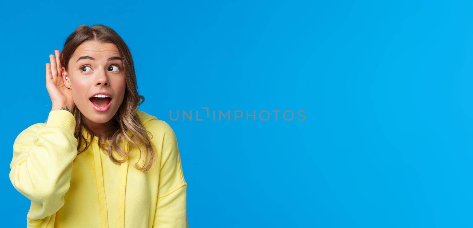 Close-up portrait of intrigued and curious blond young girl with pierced ear, gossiping, eavesdropping and listening to someone conversation interested, standing blue background by Benzoix