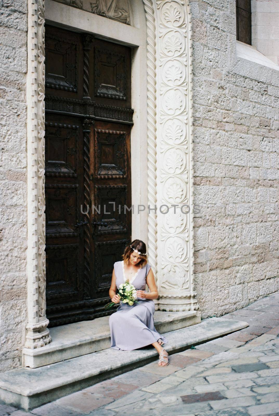 Bride with flowers in her hands sits on the steps near the wooden carved door. High quality photo