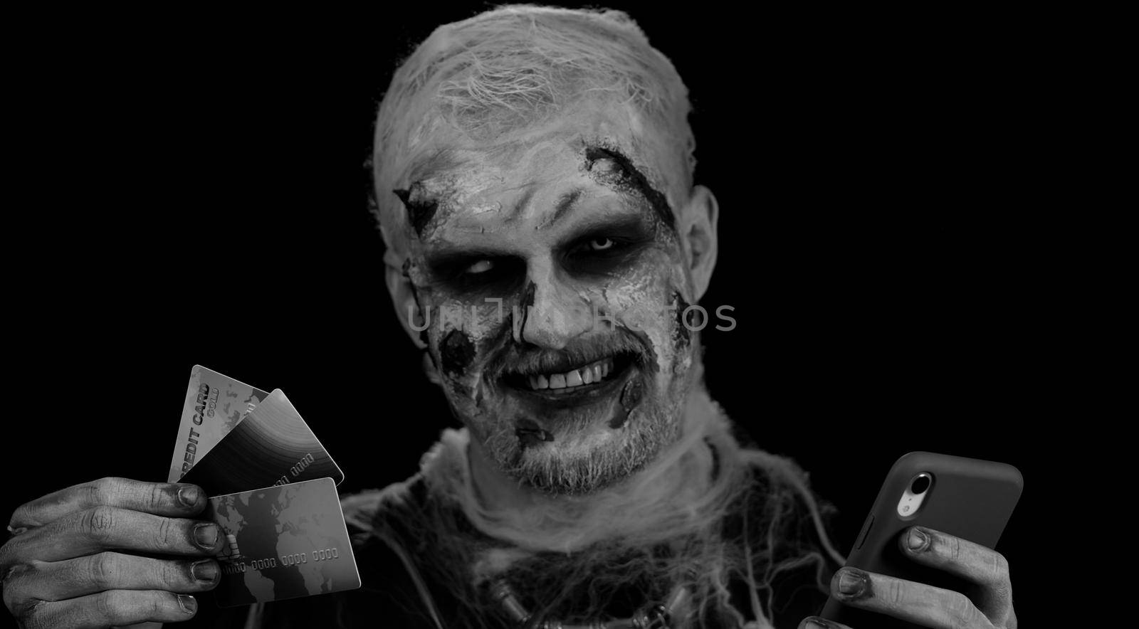 Sinister man with horrible scary Halloween zombie make-up using credit bank cards and mobile phone, transferring money purchases online shopping. Dead guy with wounded bloody scars face, black room