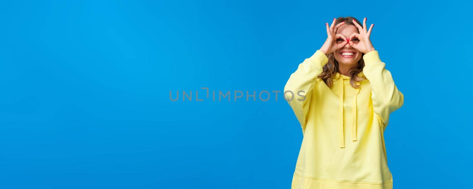 Dreamy and silly girl looking through fingers with happy playful expression, make okay gestures form glasses with hands and smiling carefree, look far away, searching something, blue background.