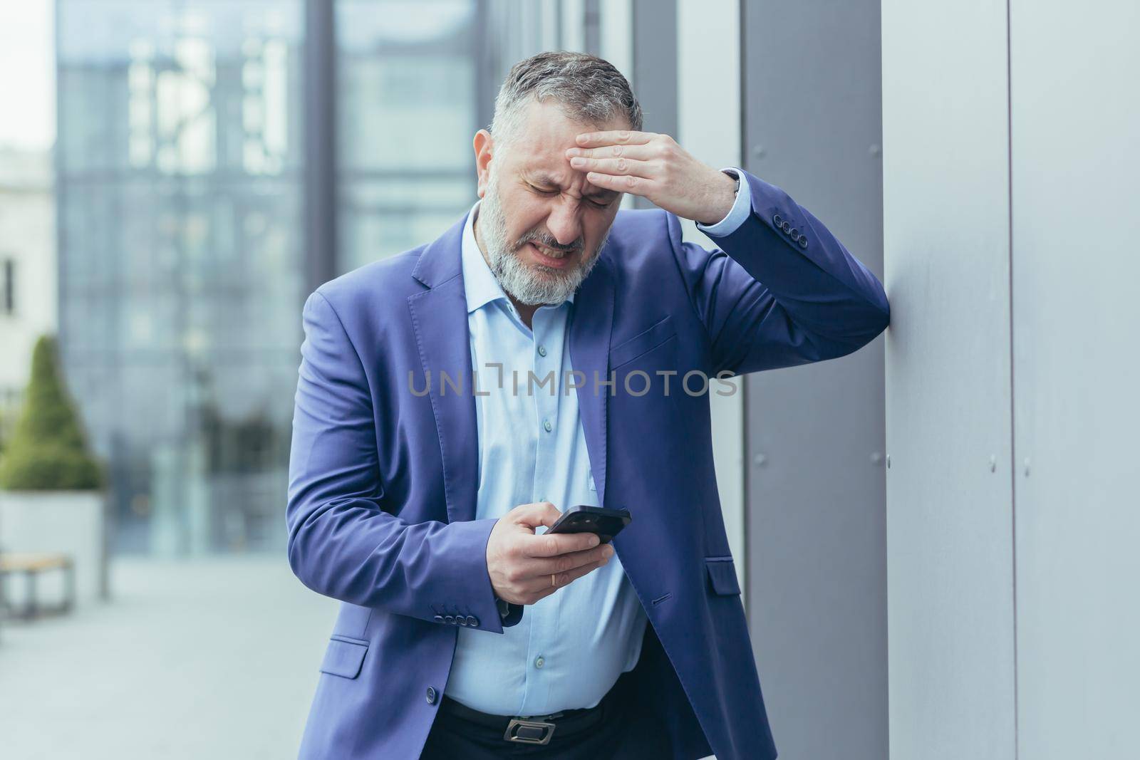 A senior man, a businessman, is standing on the street near an office center, holding his head in his hand, having a severe headache. Calls an ambulance by phone.