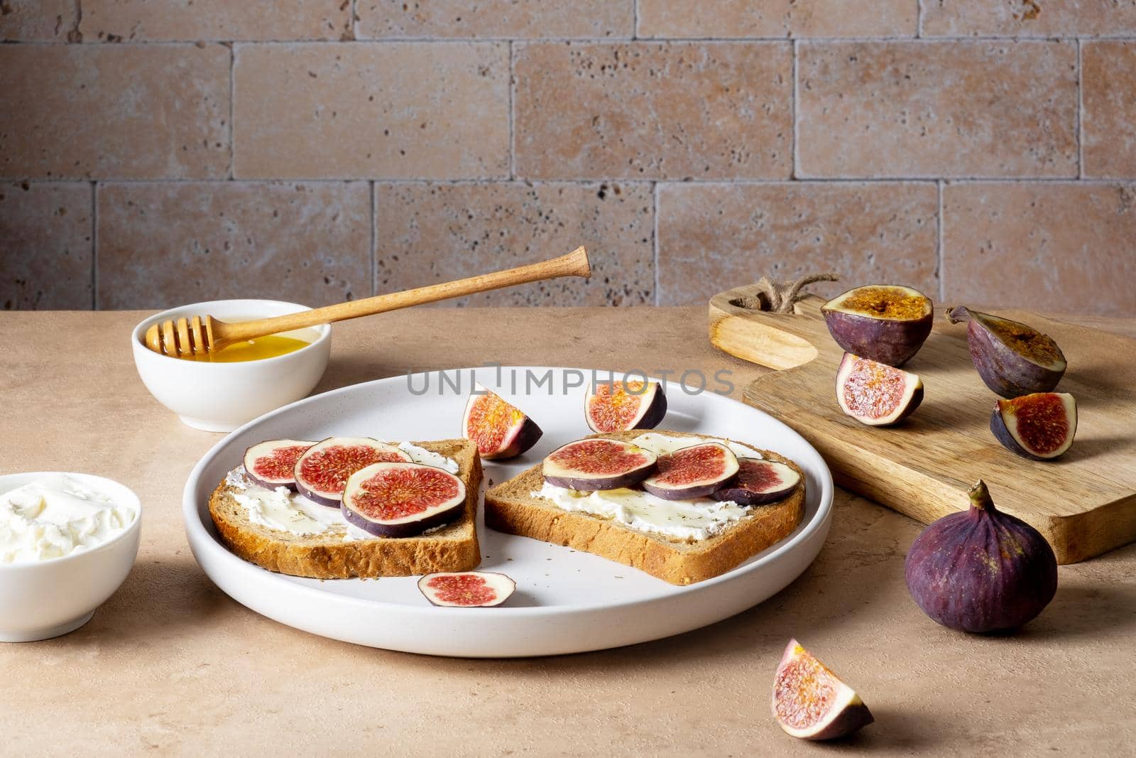 Bruschetta with figs, cream cheese and honey lie on light ceramic plate on light beige table against backdrop of masonry wall. Ingredients. Healthy food. Selective focus.