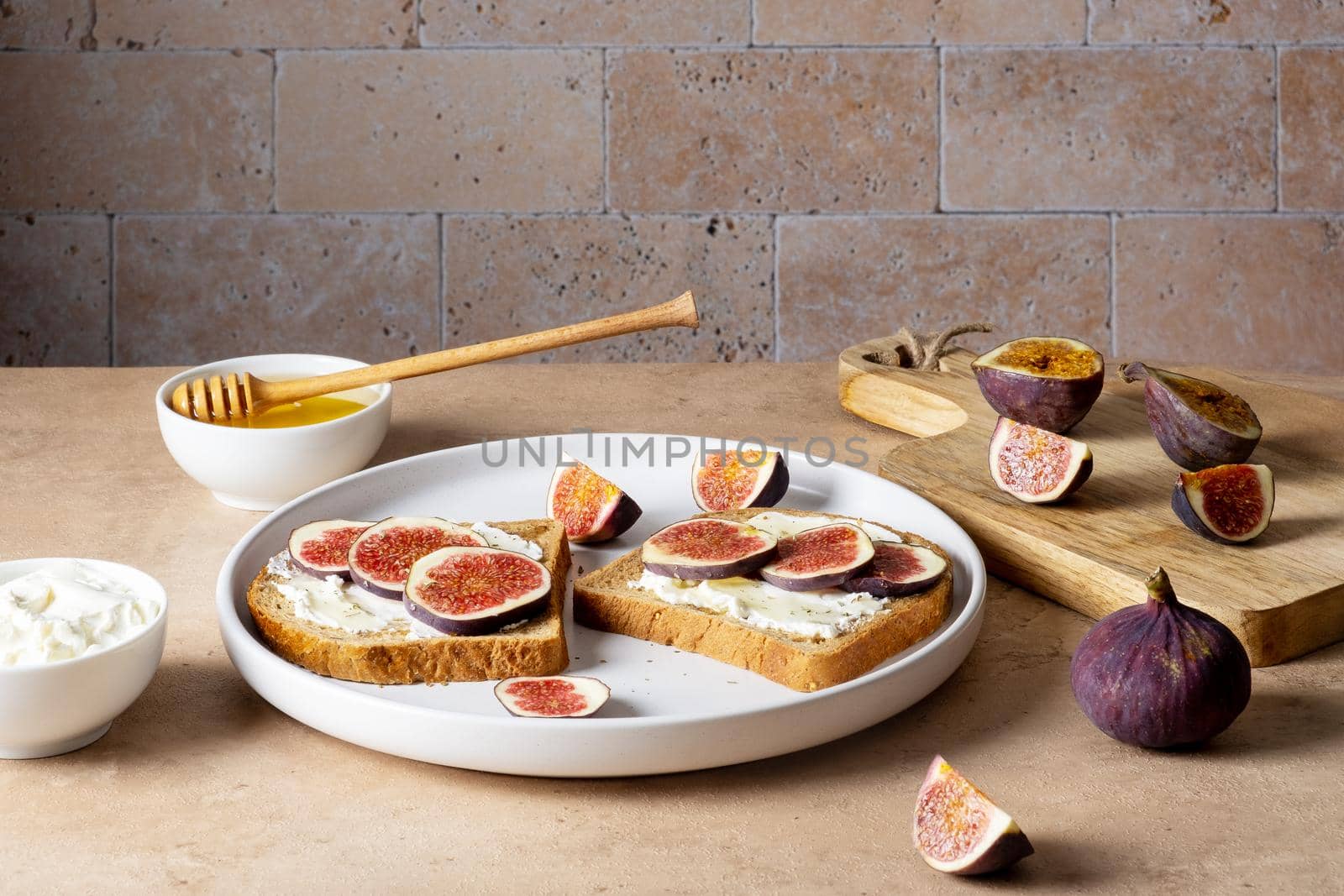 Bruschetta with figs, cream cheese and honey lie on light ceramic plate on light beige table against backdrop of masonry wall. Ingredients. Healthy food. Selective focus.