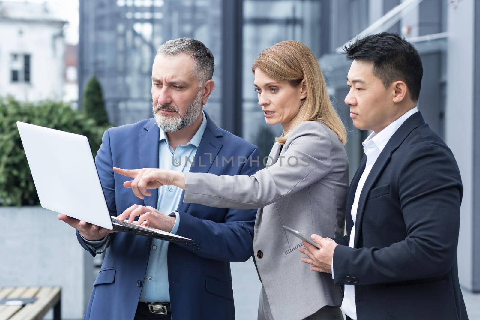Successful business team, three colleagues businessman and businesswoman outside office building discussing current plans and management, looking at laptop screen, discussing and consulting