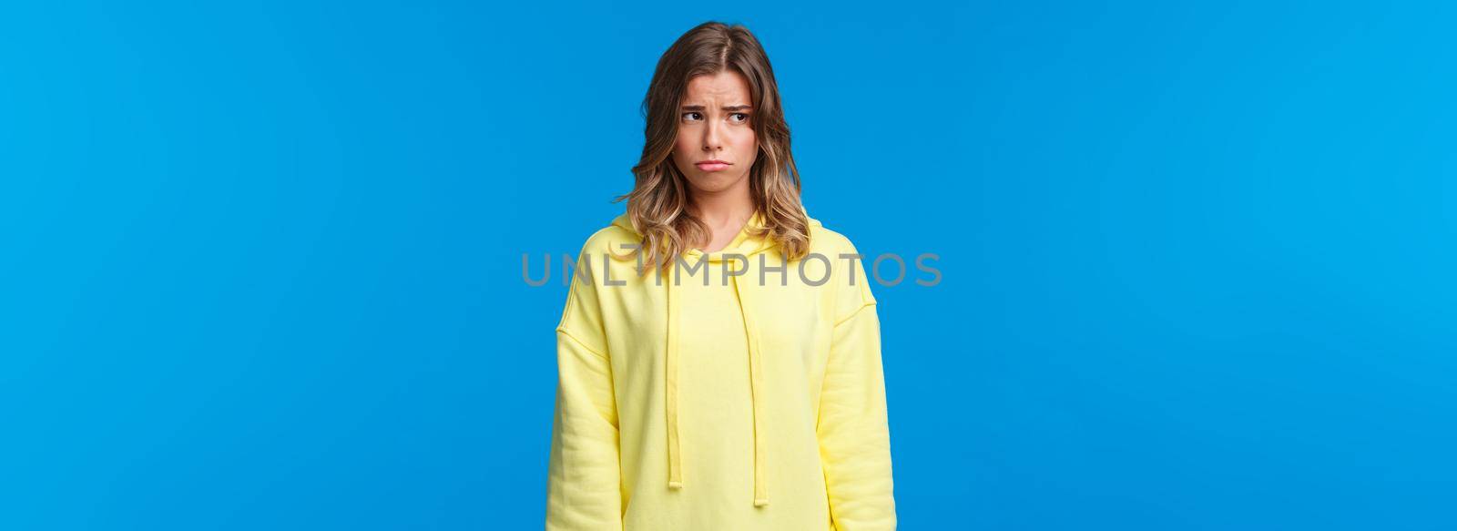 Gloomy and lonely say caucasian girl looking away, sobbing childish and sulking as feeling uneasy, being let down, having bad day at university, standing upset over blue background.