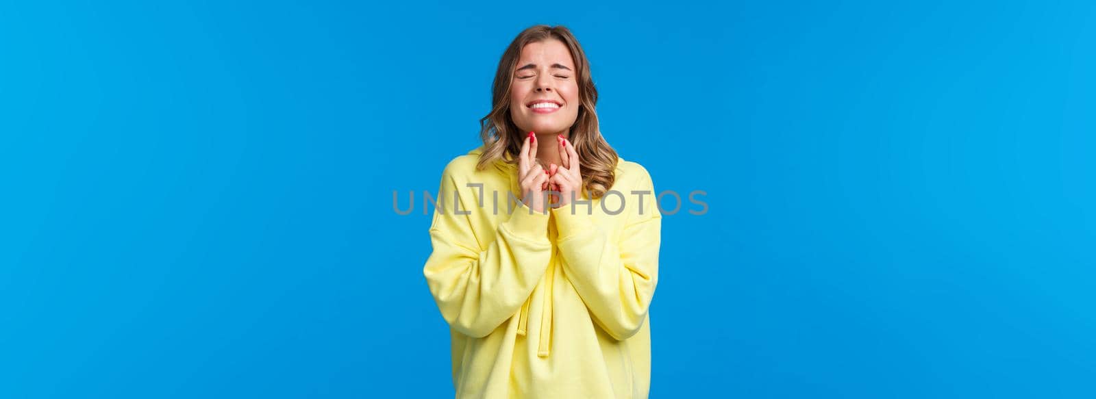 Wishing all well. Hopeful optimistic blond girl in yellow hoodie praying god make wish come true, cross fingers good luck pleading with closed eyes, supplicating, stand blue background.