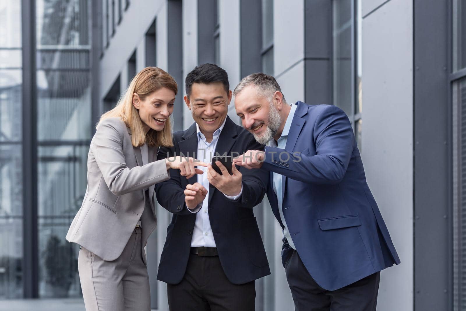 Diverse business group, three male and female workers outside office building cheerfully smiling and happy watching video on phone