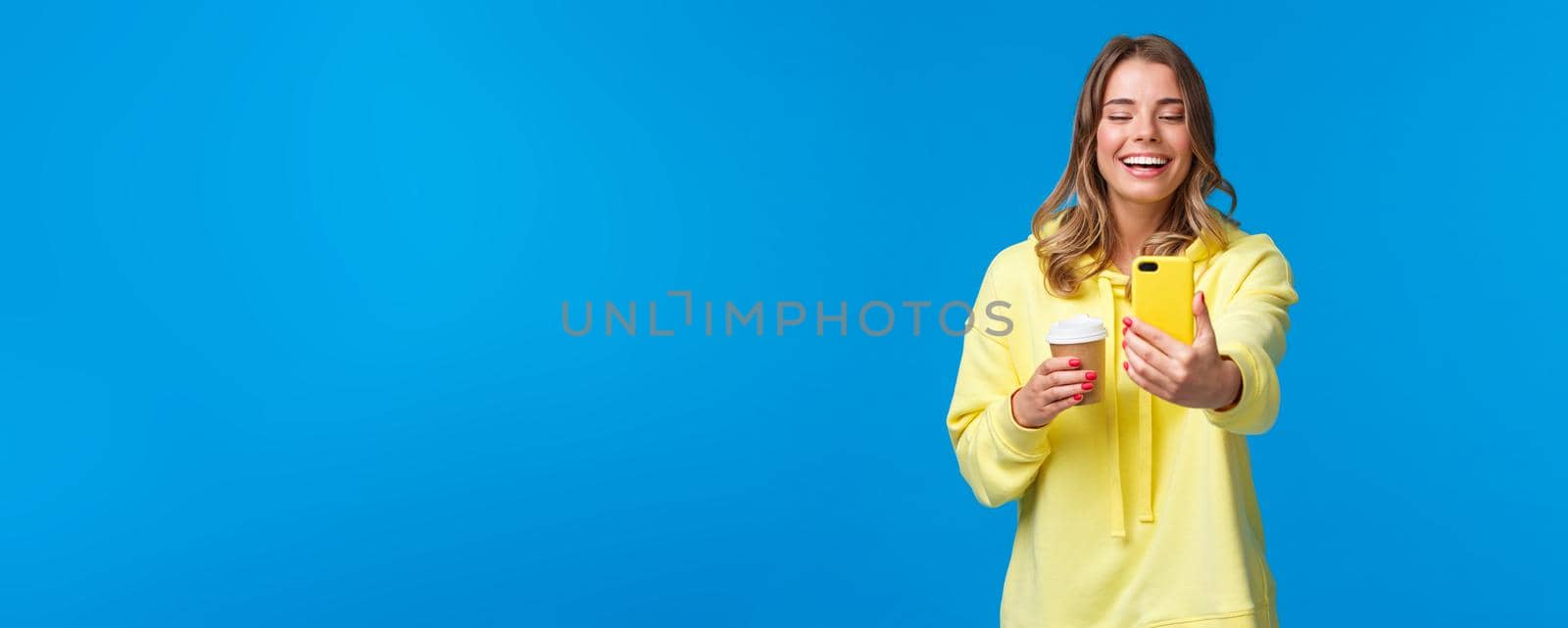 Cheerful smiling blond caucasian female blogger record video or take selfie on her smartphone, laughing and grinning as holding take-away cup of coffee, stand blue background.