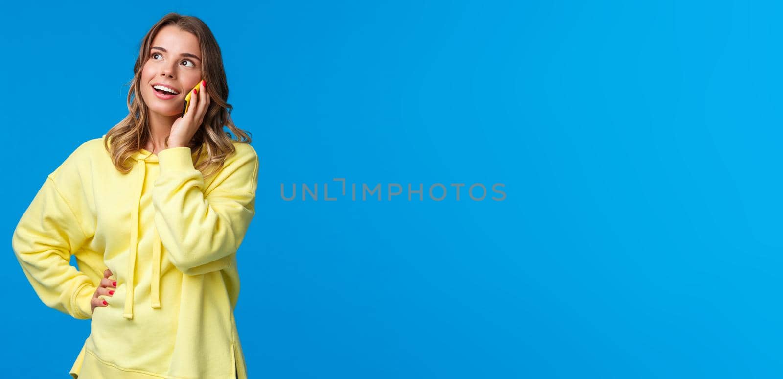 Friendly carefree good-looking blond woman talking on phone, holding mobile phone near ear and look up with pleased smile, thinking, making order or calling friend, blue background.