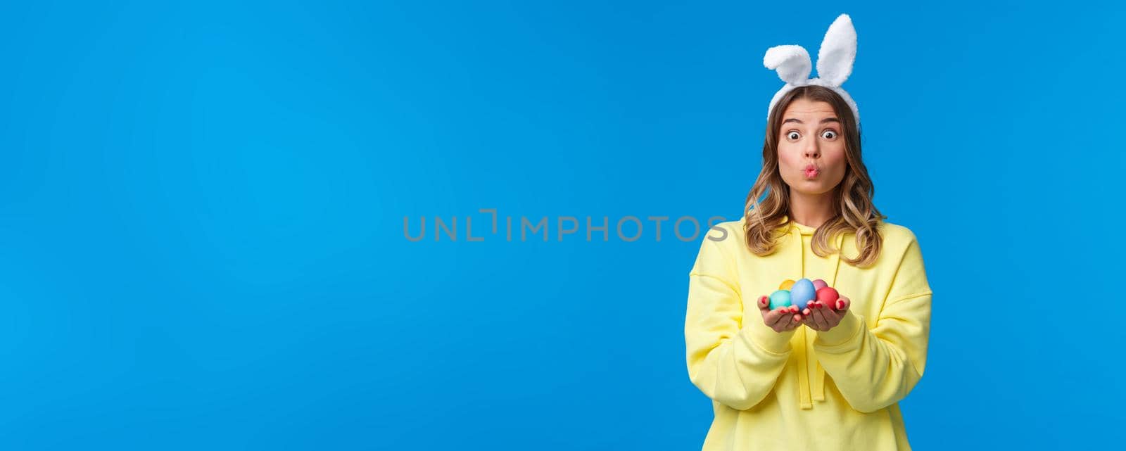 Holidays, traditions and celebration concept. Silly cute caucasian blond girl present you Easter eggs, painted it for holiday, show mwah kiss expression and wear rabbit ears, blue background by Benzoix