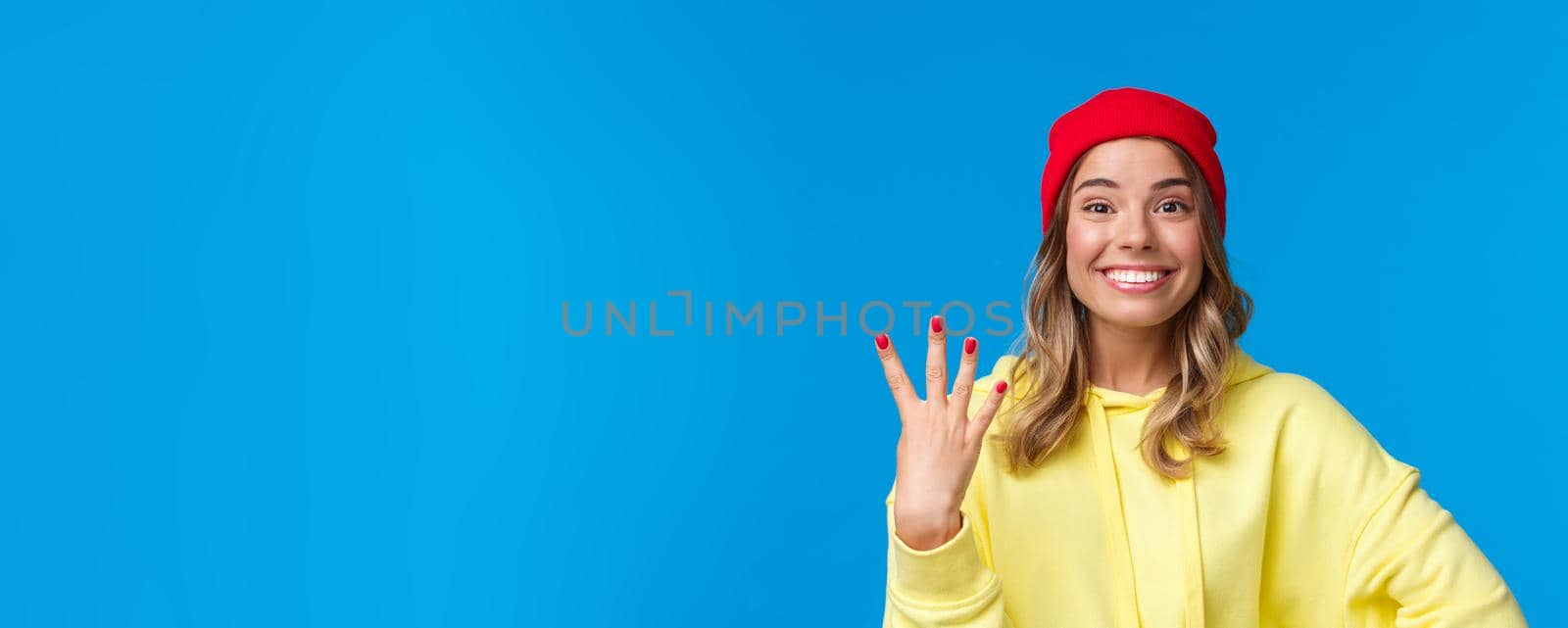 Happy smiling female student in hipster red beanie, showing number four, fourth and grinning at camera optimistic, make reservation or order, standing blue background upbeat. Copy space