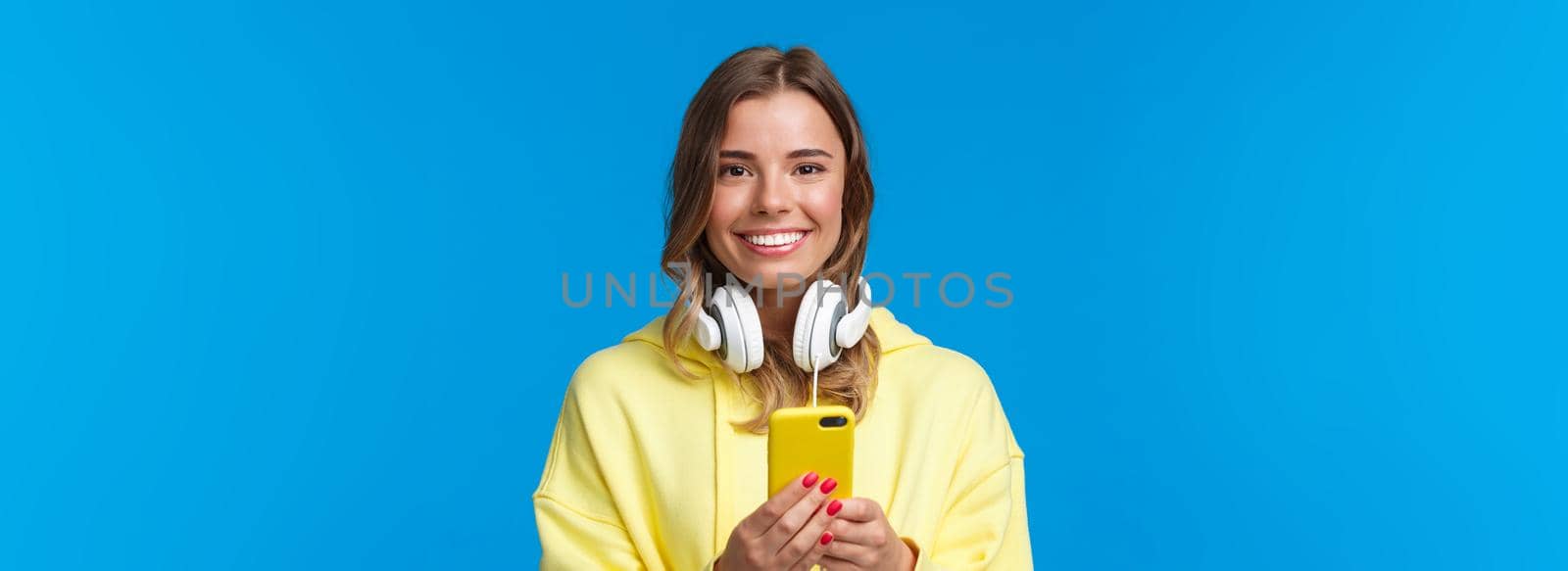 Close-up portrait of young blond european hipster girl with blond short hair and headphones over neck, holding smartphone, texting message, use mobile phone, blue background by Benzoix