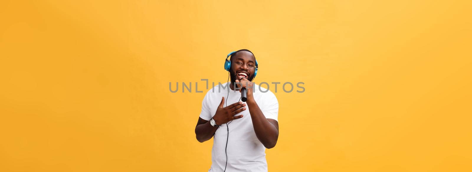 Portrait of cheerful positive chic. handsome african man holding microphone and having headphones on head listening music singing song enjoying weekend vacation isolated on yellow background.