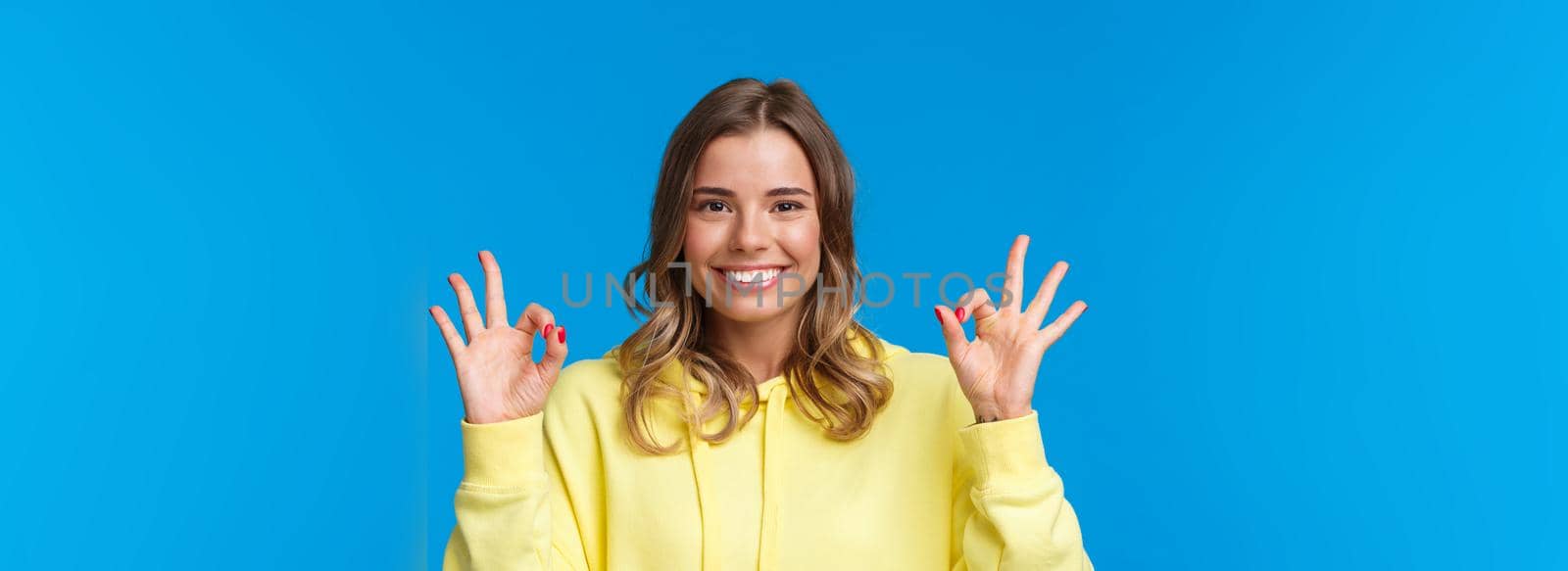 Close-up portrait happy confident young blond girl assure all okay, guarantee plan went well, smiling and show ok gesture in approval, confirmation or like, stand blue background. Copy space