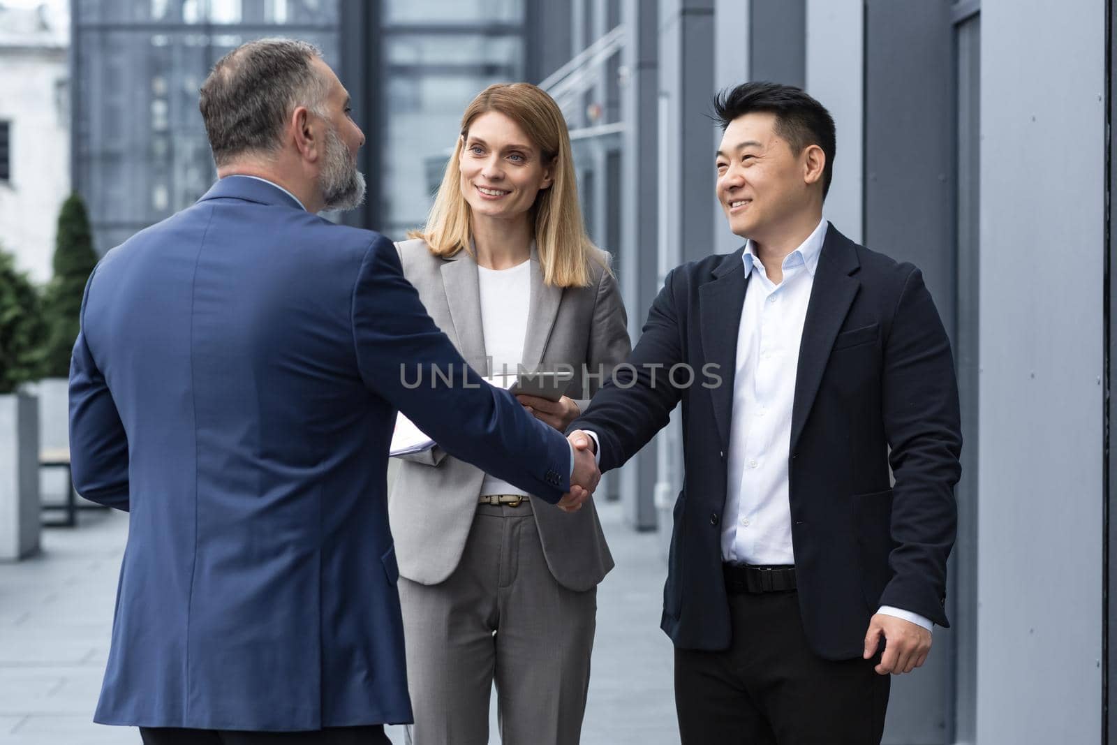 Meeting of three successful business people, diverse dream team man and woman outside office building, greeting and shaking hands, experienced professionals specialists in business suits talking