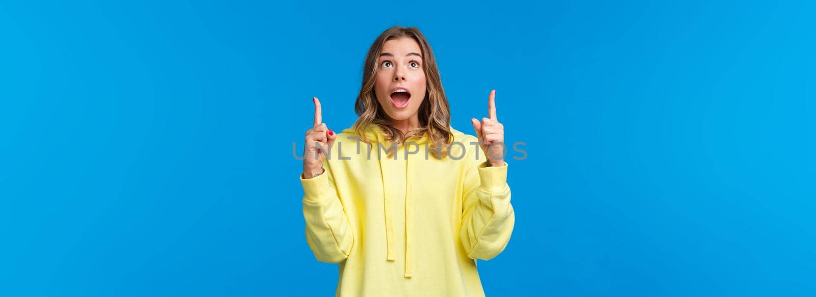 Fascinated and amused young blond girl drop jaw and staring speechless, pointing fingers up at something incredible and awesome, watching amazing performance, stand blue background by Benzoix