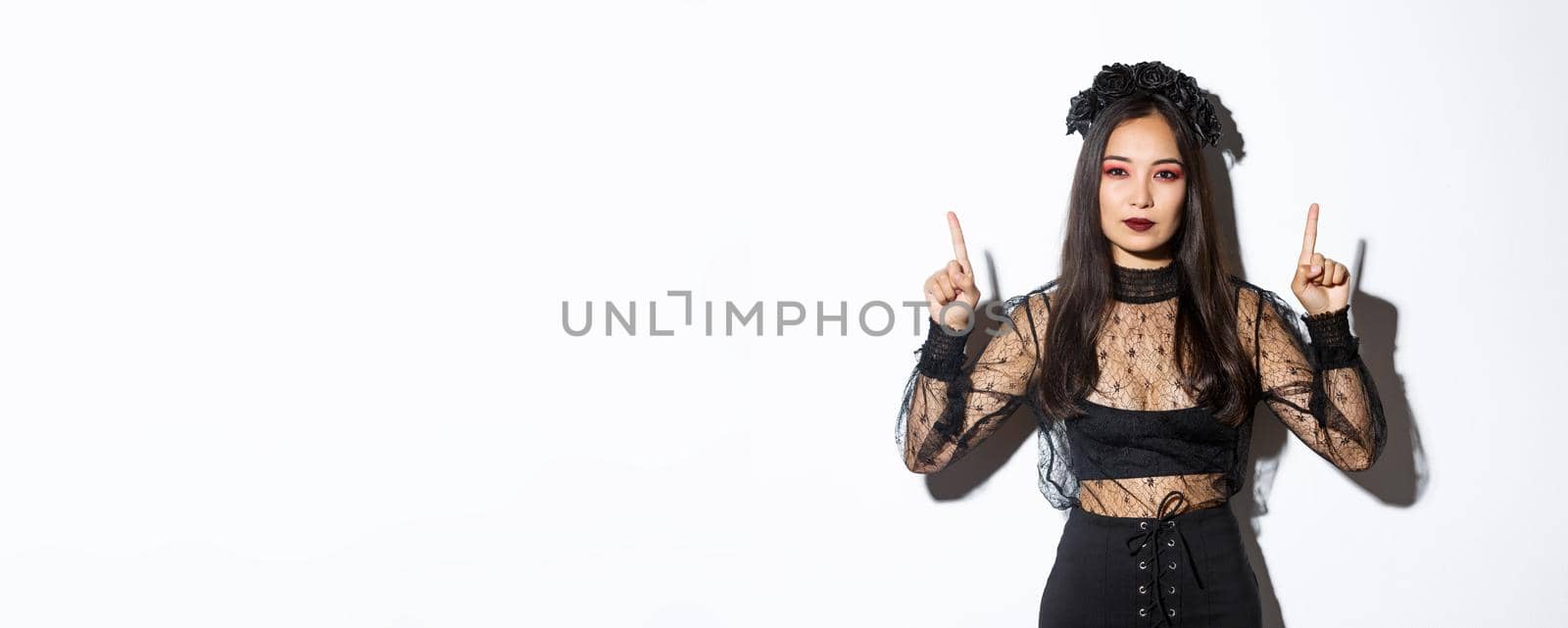 Sassy beautiful asian woman in black gothic dress, wearing witch costume for halloween and pointing fingers up, showing your logo or banner on empty white background, white background.