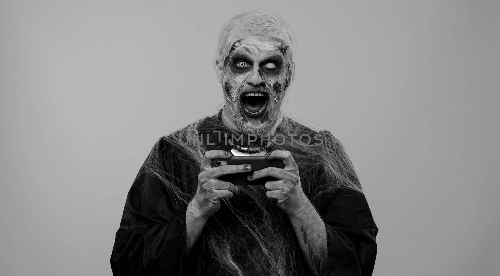 Spooky one man with horrible scary Halloween zombie makeup enthusiastically playing racing drive video games on mobile phone. Dead guy with wounded bloody scars face isolated against gray background