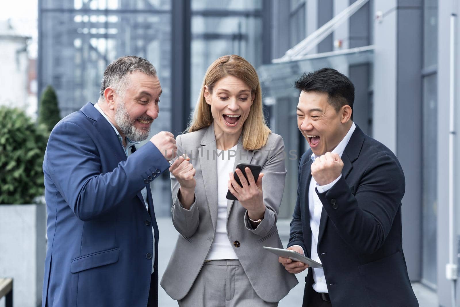 group of colleagues employees read good news looking at smartphone. Happy team rejoices about successful investment while using, browsing mobile phone. Standing outside near office building.