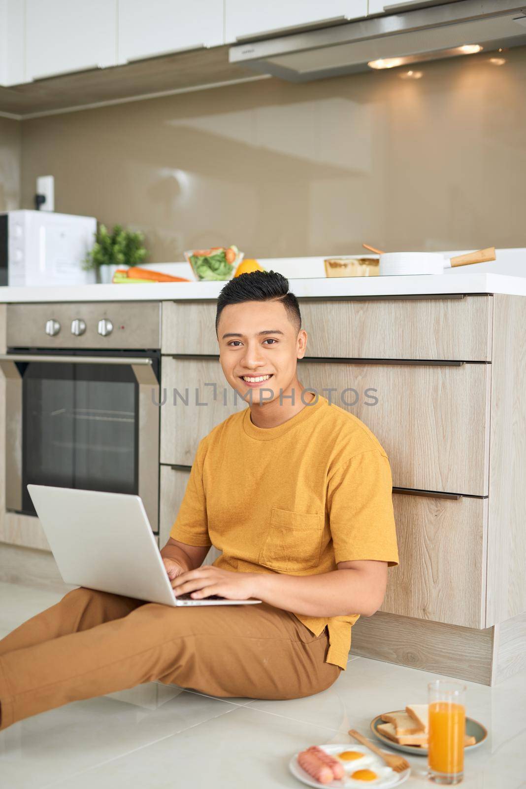 Young business man is smiling, eating breakfast and working on laptop in bright kitchen.