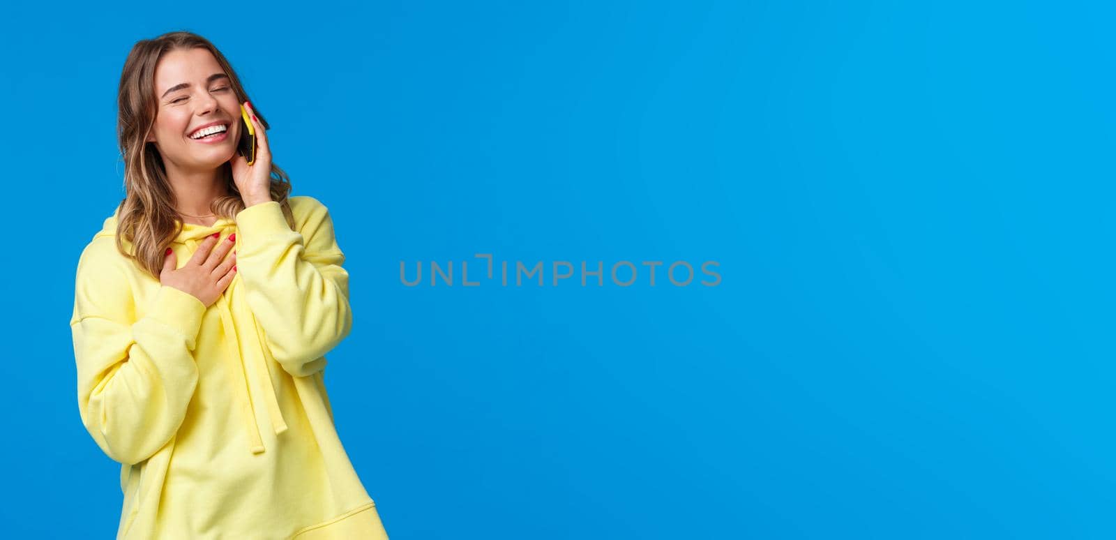 Waist-up portrait of joyful smiling young woman having funny conversation on phone, holding smartphone near ear, close eyes and laughing as touch chest, standing blue background by Benzoix