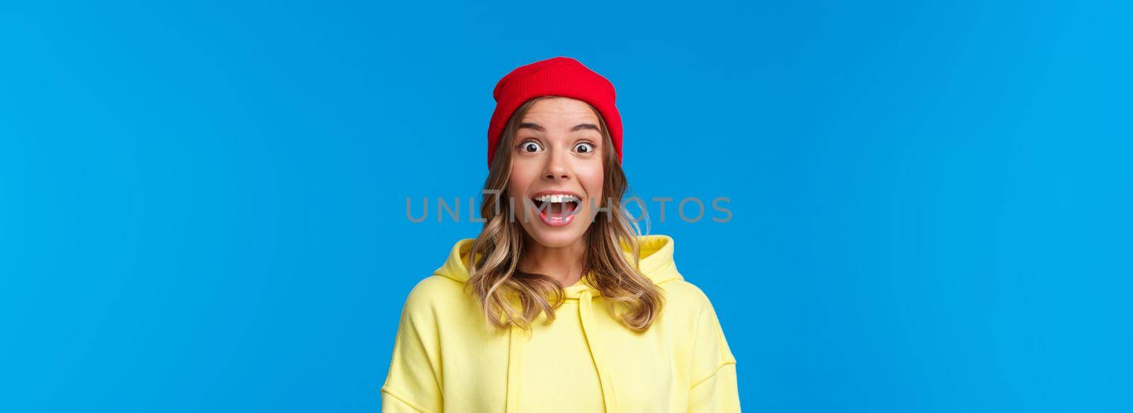 Close-up portrait of excited and intrigued young hipster girl in red beanie and yellow hoodie, gasping amazed look camera wondered and entertained, standing blue background by Benzoix