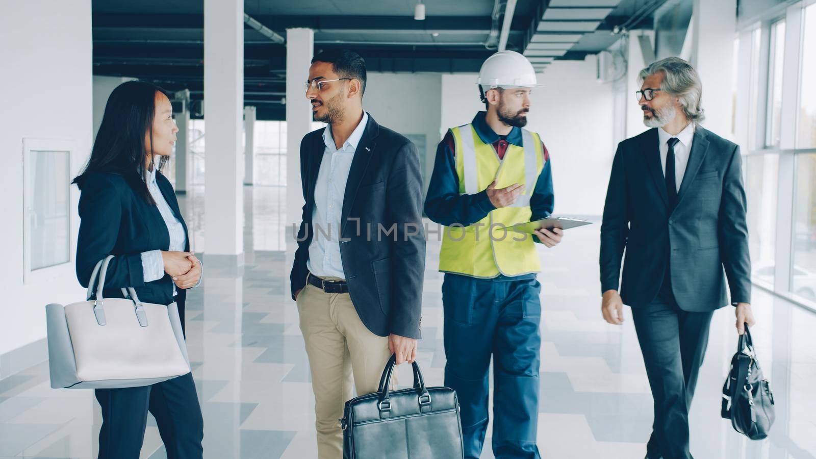 Multi-ethnic group of investors shaking hands and talking to builder in uniform by silverkblack