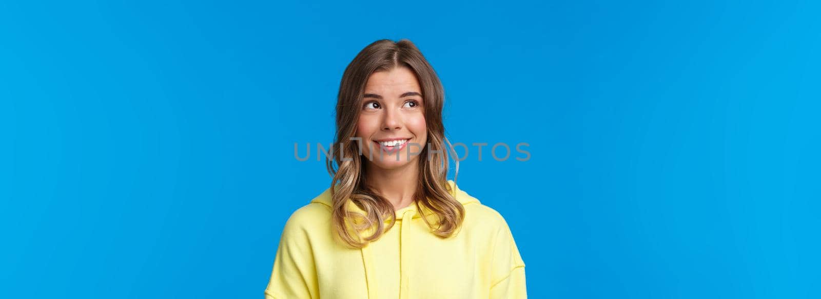 Close-up portrait of attractive european blond female with short hair, wear yellow hoodie, looking dreamy and silly upper left corner, smiling with white beaming smile, standing blue background.