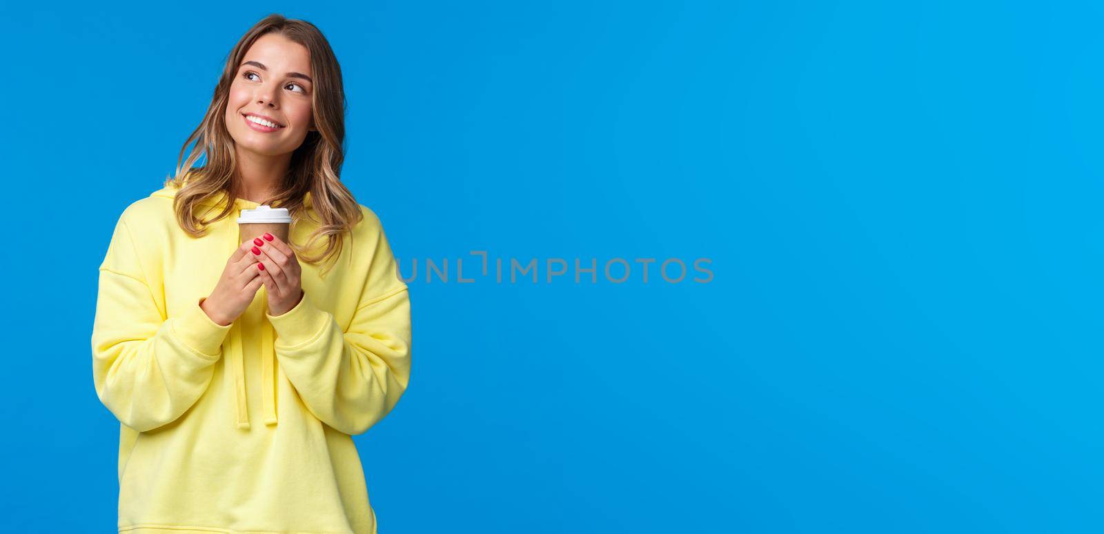 Dreamy cute blond european girl in yellow hoodie looking up romantically and thoughtful, smiling to herself as imaging something kawaii holding paper cup drinking coffee, blue background.