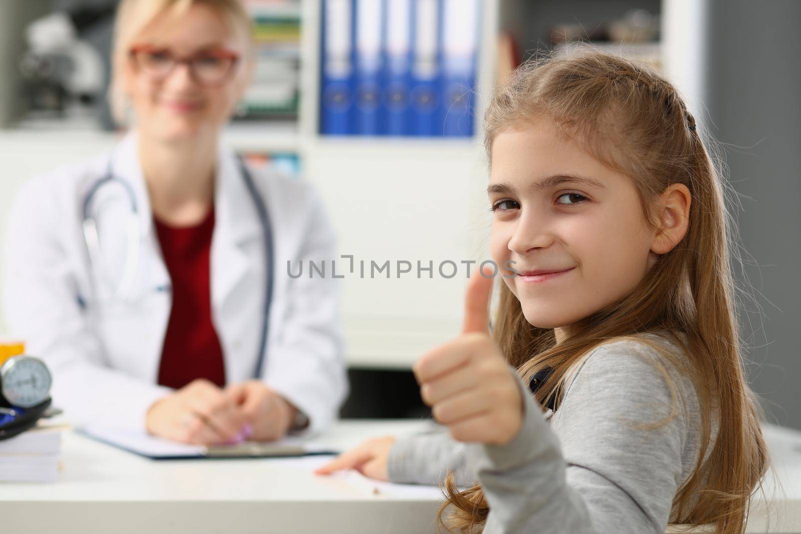 Beautiful little girl shows thumbs up. and doctor sits in background. Health insurance and examination of children