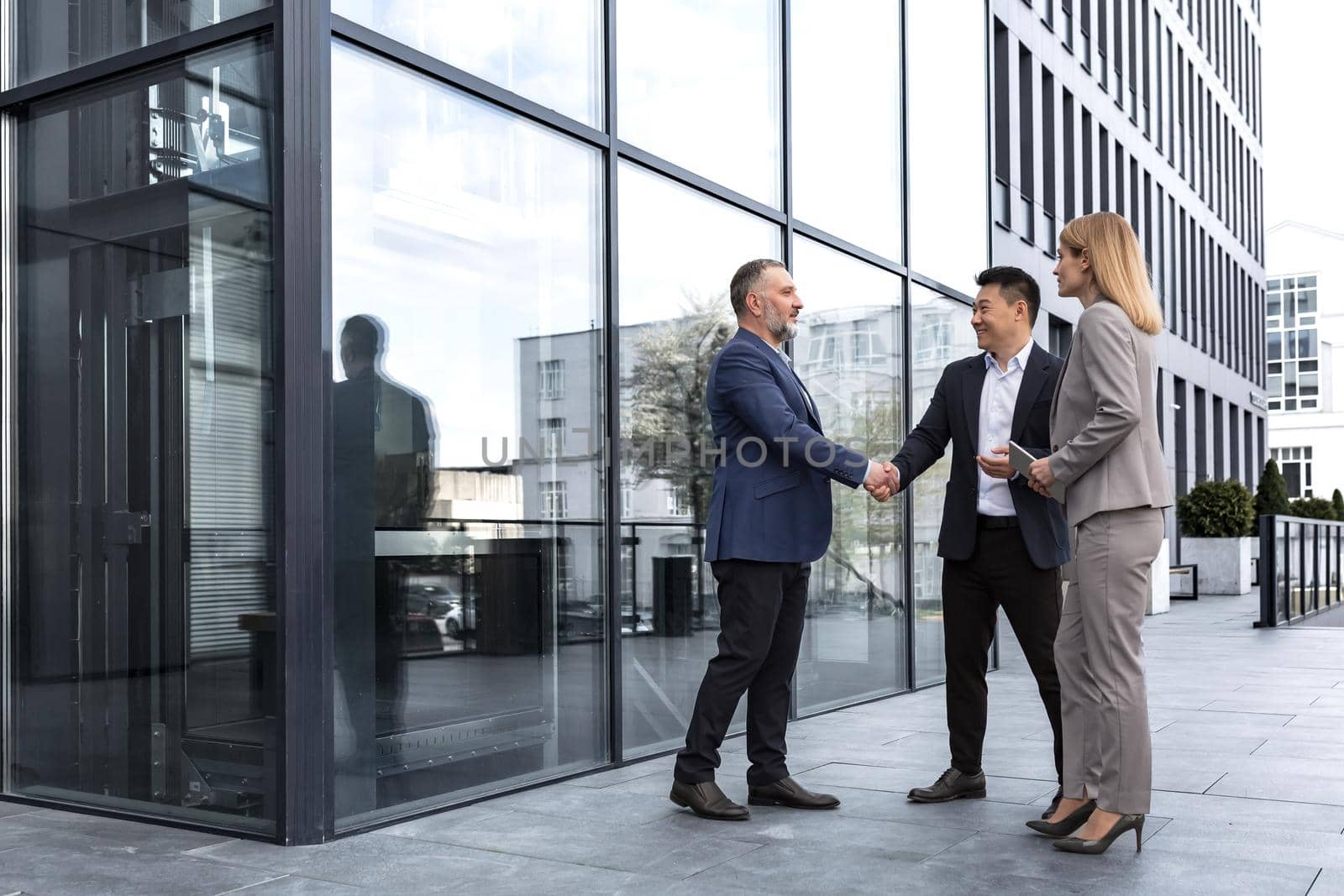 Meeting of three successful business people, diverse dream team man and woman outside office building, greeting and shaking hands, experienced professionals specialists in business suits talking