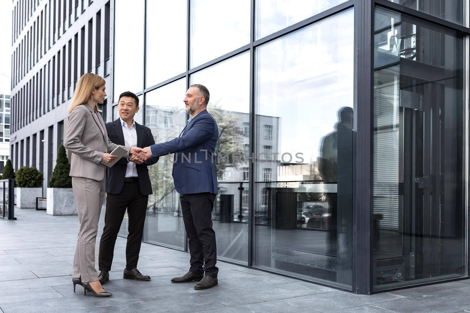 Meeting of three colleagues from outside the office building, experienced and mature IT specialists, greeting and shaking hands, business persons in business suits, diverse group of people