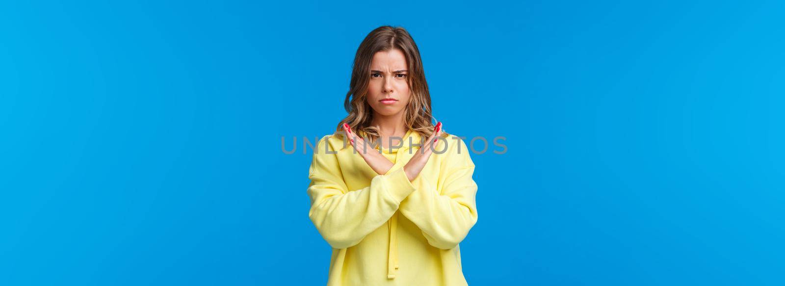 Stop right there, no means no. Serious-looking confident blond woman in yellow hoodie making cross on chest, enough and decline gesture, demand quit it, stand blue background.