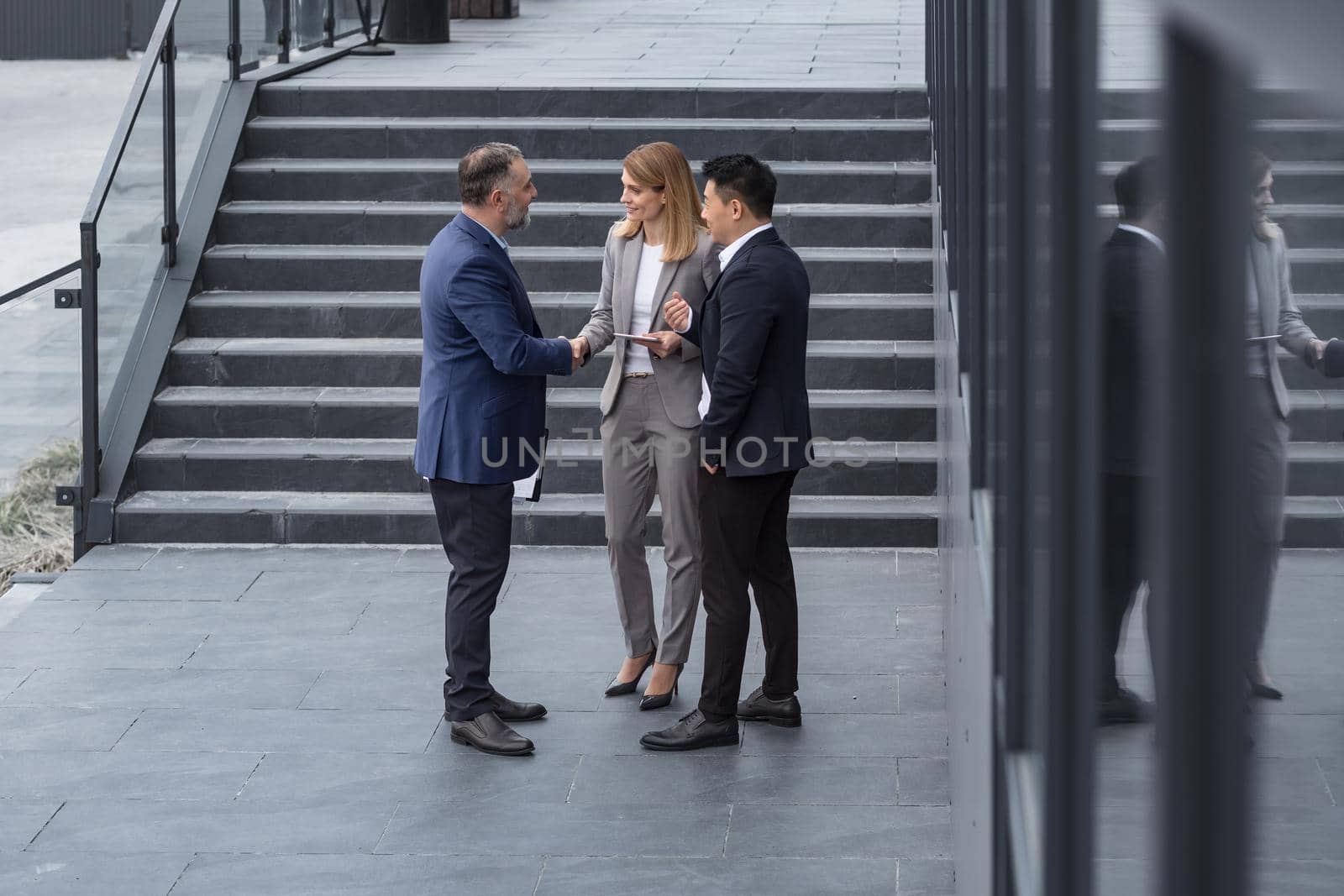 Meeting of three successful business people, diverse dream team man and woman outside office building, greeting and shaking hands, experienced professionals specialists in business suits talking