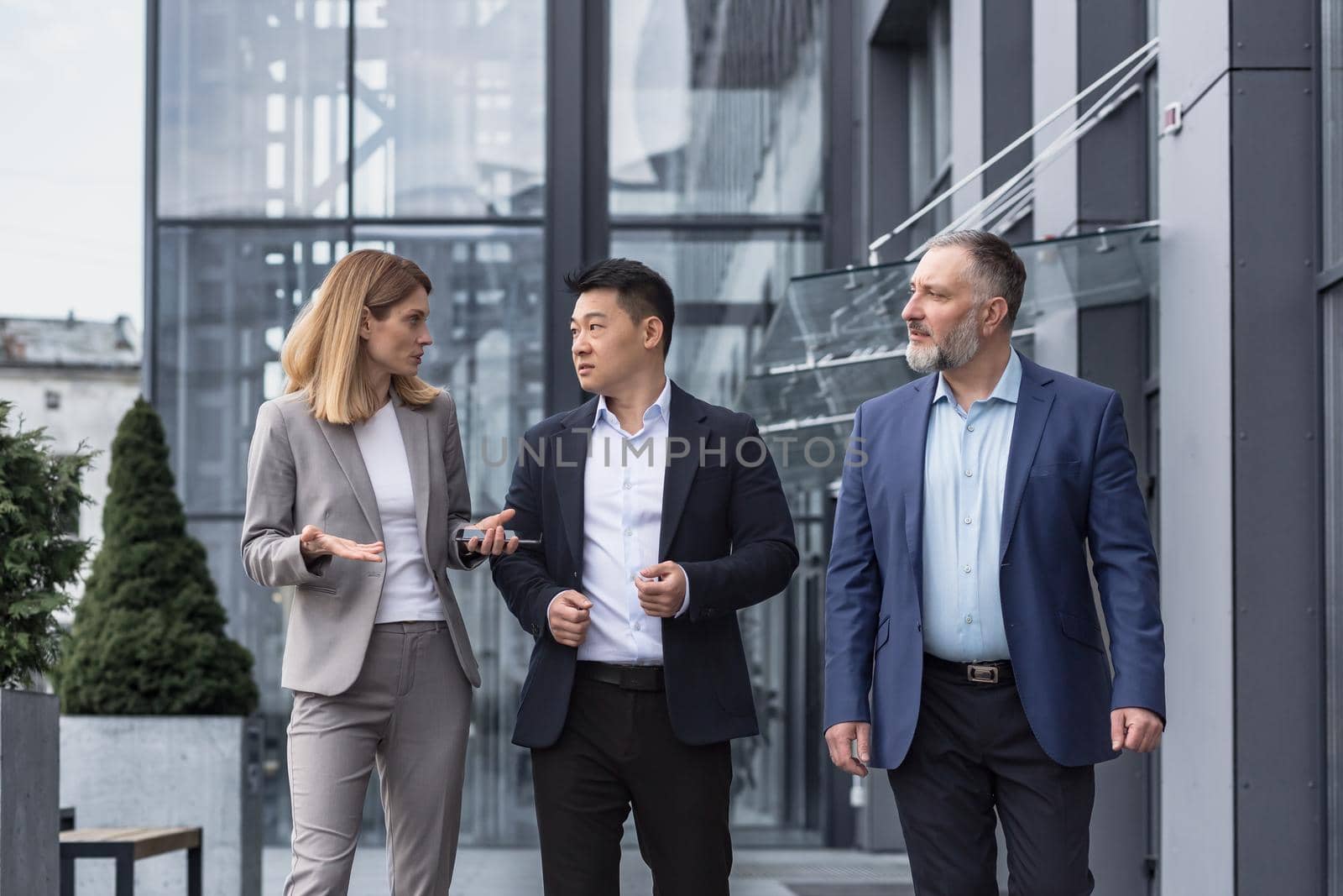 Three diverse business people walking and talking focused and thoughtful seriously outside office building, male and female, discussing plans and work project