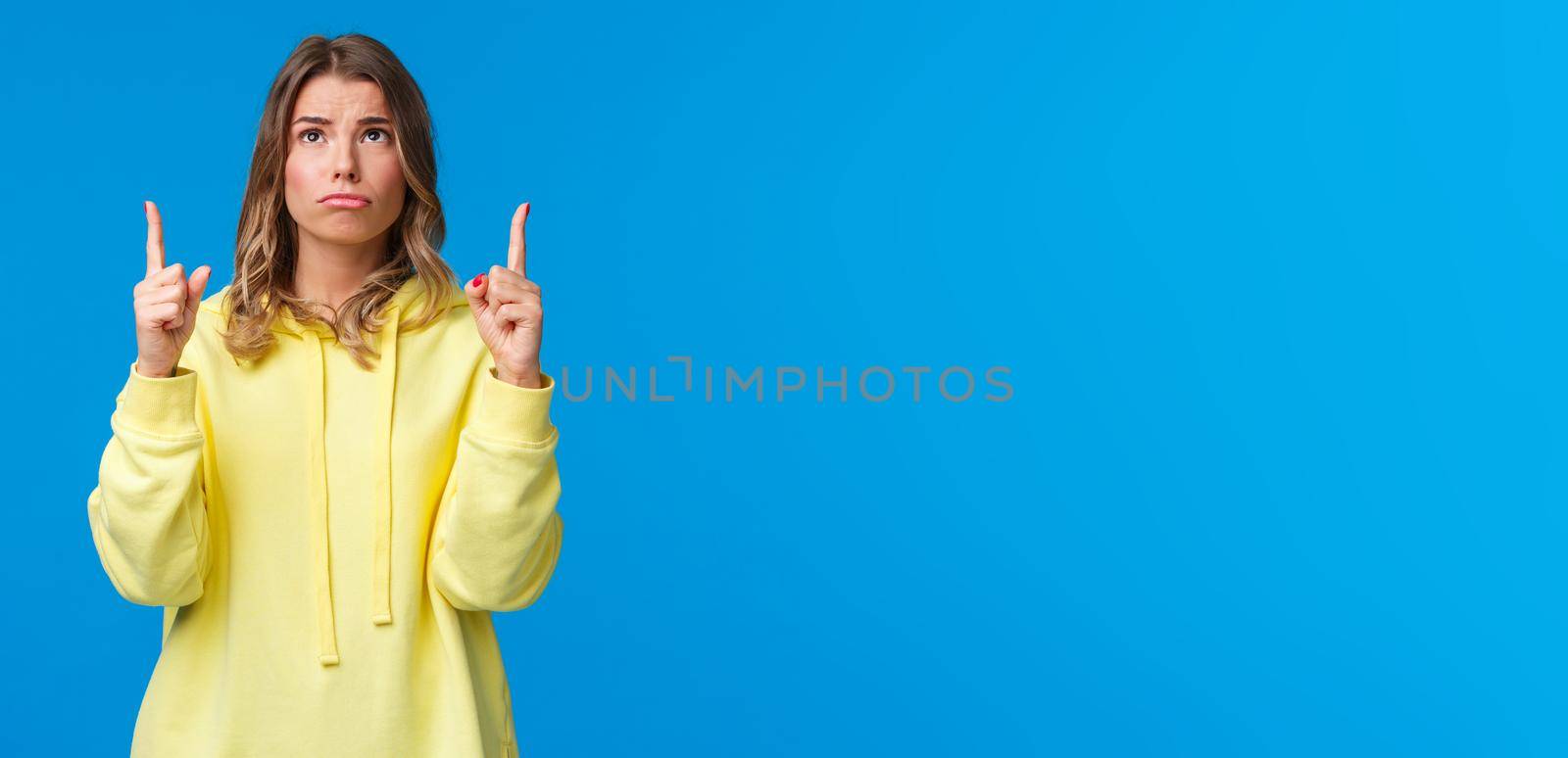 Jealous and gloomy cute blond female student feel regret not having something she desires, sobbing frowning sad as looking pointing up, standing over blue background.