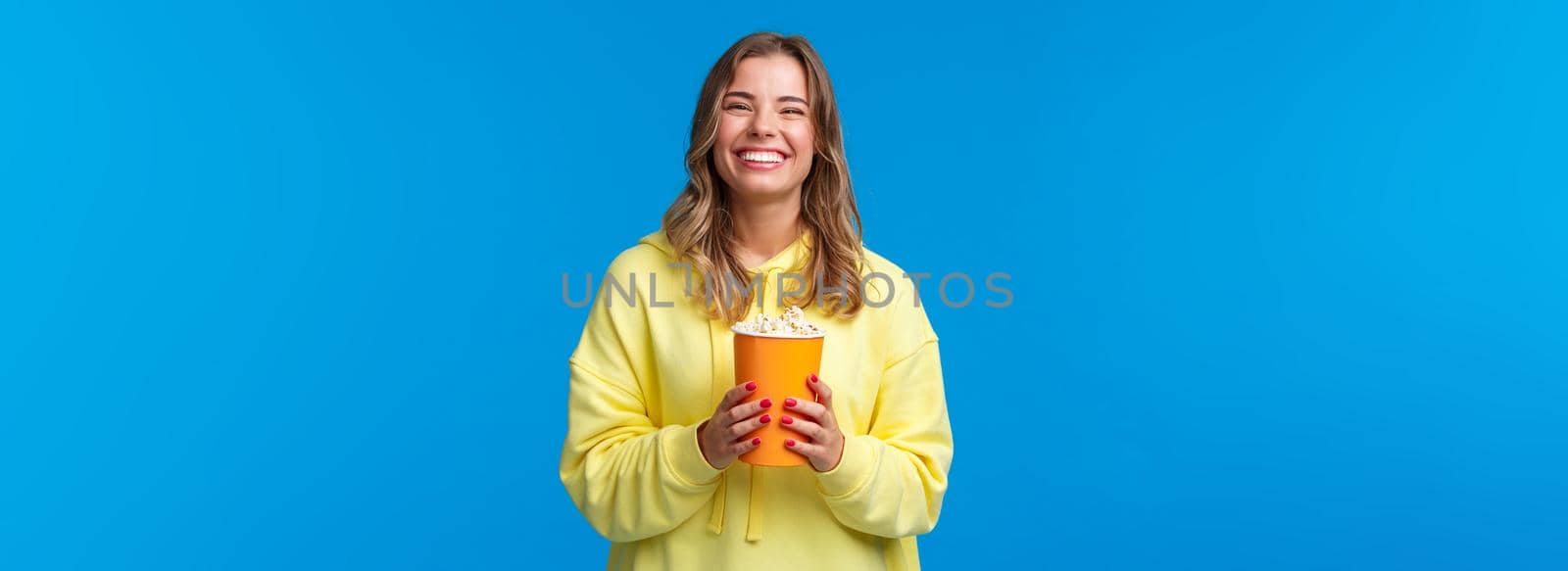 Leisure, fun and youth concept. Friendly happy blond european woman enjoying weekends, eating popcorn and watching comedy, visit cinema to see new movie, stand blue background.