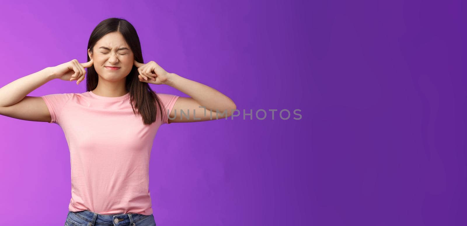 Displeased annoyed asian brunette woman fed up hearing loud parents yell, close eyes grimacing, plug ears fingers, disturbed bothering sound, stand purple background unsatisfied. Copy space