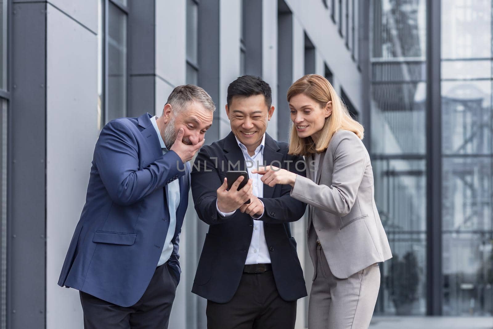 Diverse business group, three male and female workers outside office building cheerfully smiling and happy watching video on phone