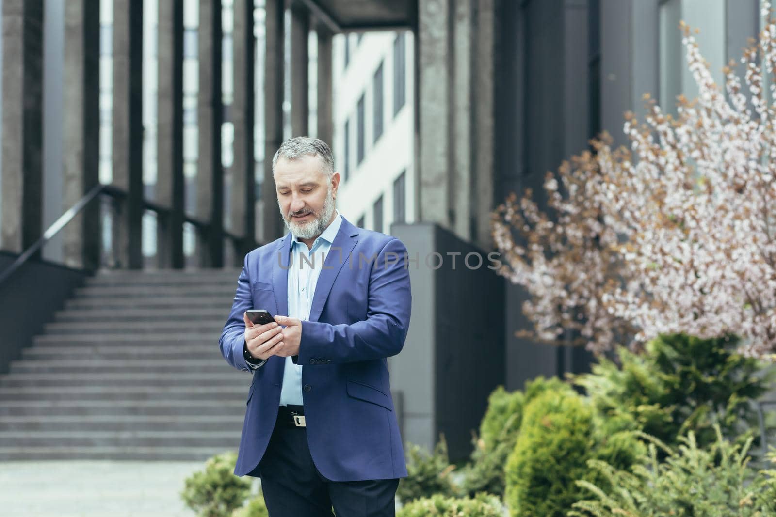 Senior gray-haired man, a businessman in a suit, stands near the modern office center and calls a taxi from the phone, typing the number, waits, is in a hurry to meet.