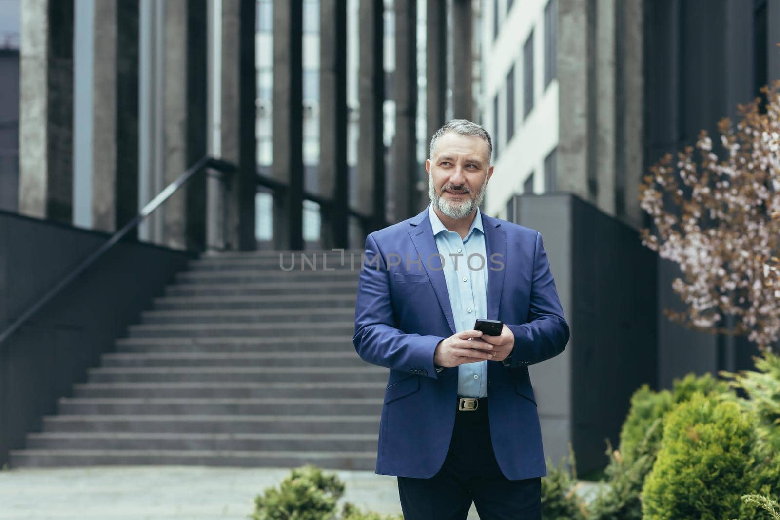 An older gray-haired man, a businessman in a suit, is traveling to another city, to another country. He is holding the phone in his hands, looking for directions on GPS. Walks near a modern building.