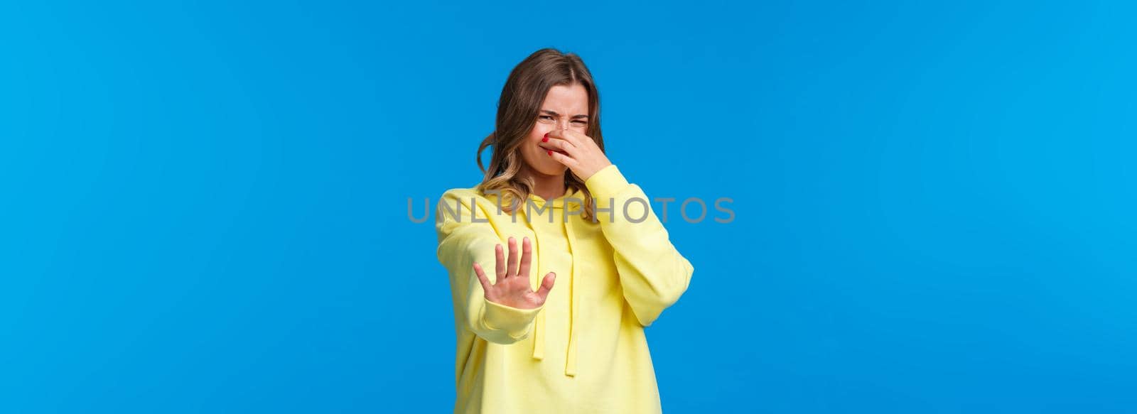 Oh gosh take this away from my face. Bothered and disturbed young blond woman close her nose and grimacing from awful nasty smell, pulling hand in stop refusal gesture, blue background by Benzoix