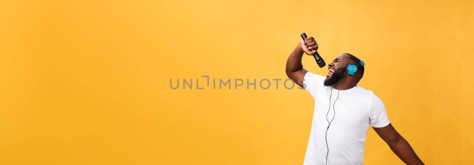 Portrait of cheerful positive chic handsome african man holding microphone and having headphones on head listening music singing song enjoying weekend vacation isolated on yellow background by Benzoix