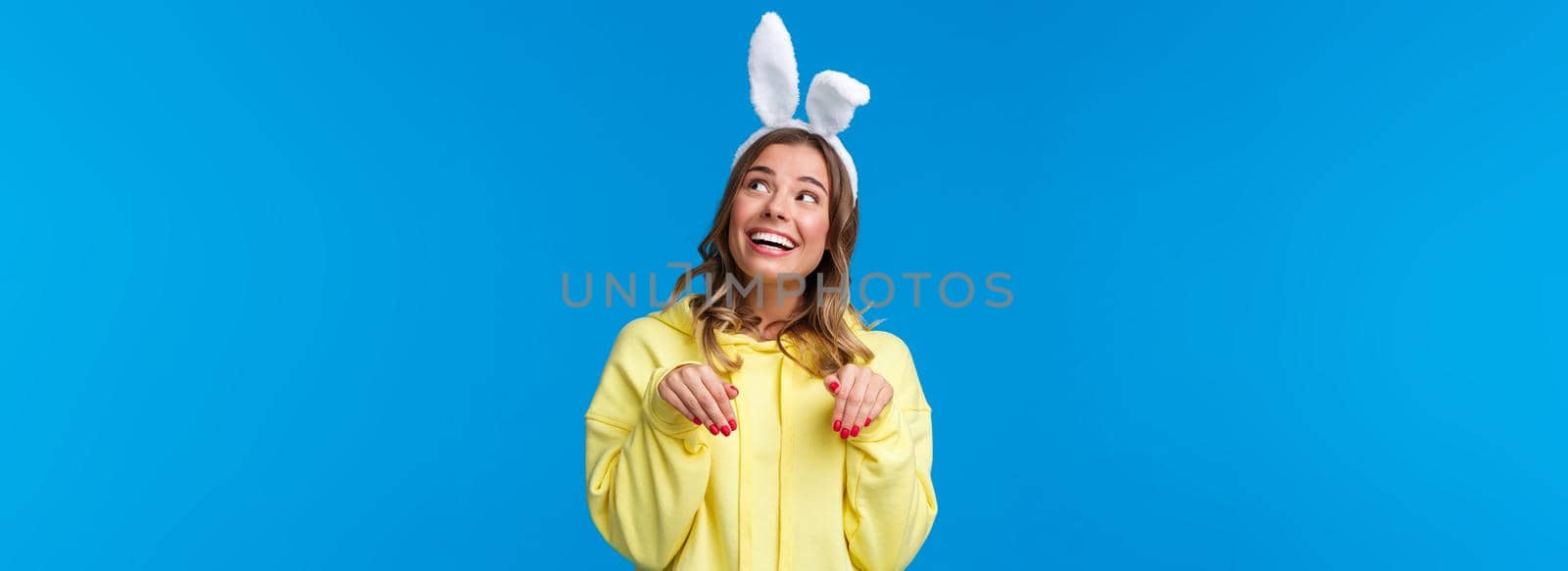 Holidays, traditions and celebration concept. Dreamy and cute young woman smiling lovely with rabbit ears, look upbeat upper right corner grinning make paws gesture, blue background.