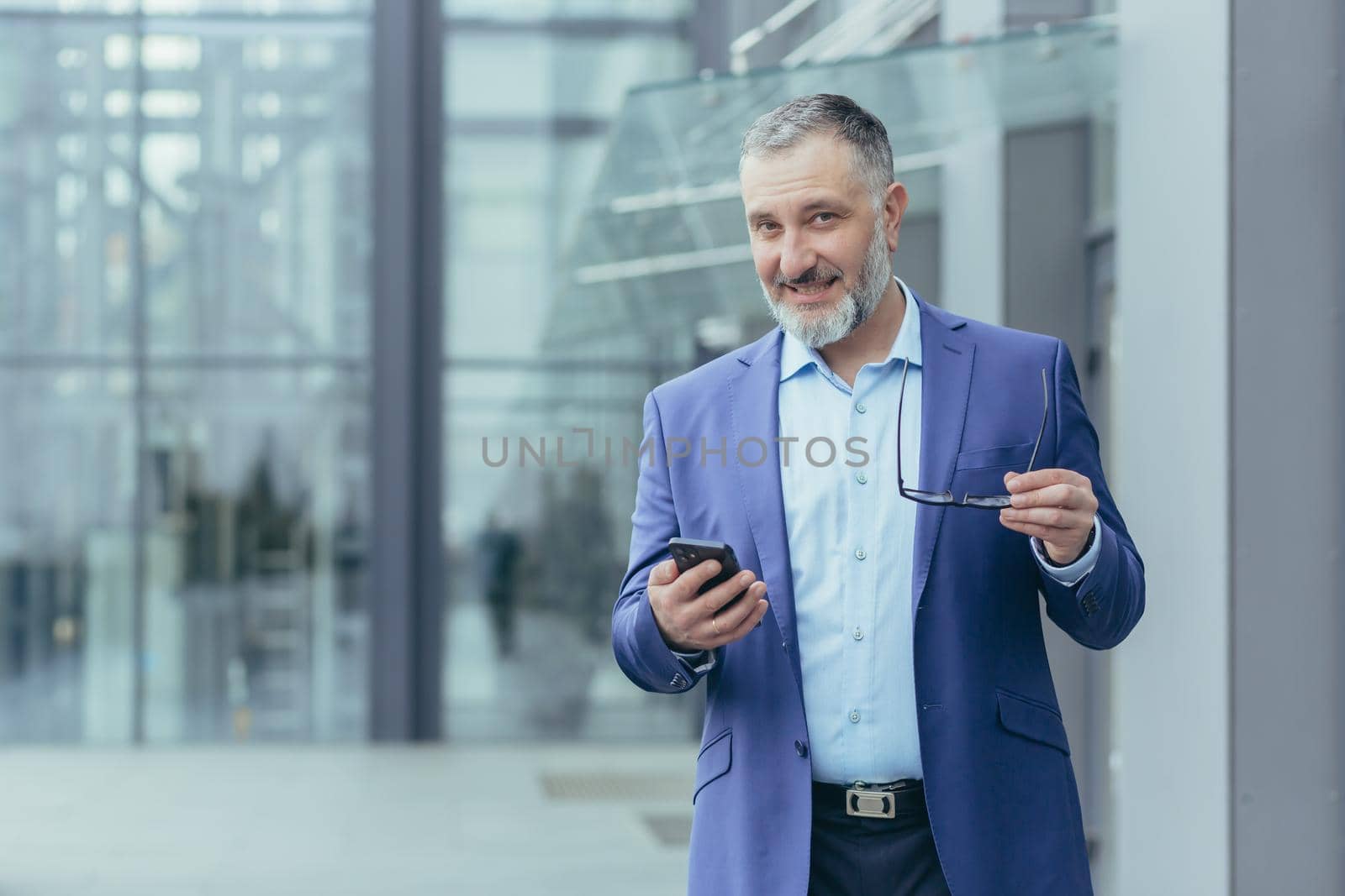 Portrait senior handsome gray-haired male teacher, professor, lecturer stands in a suit in a modern campus, holds a phone and glasses. He looks at the camera, smiles.