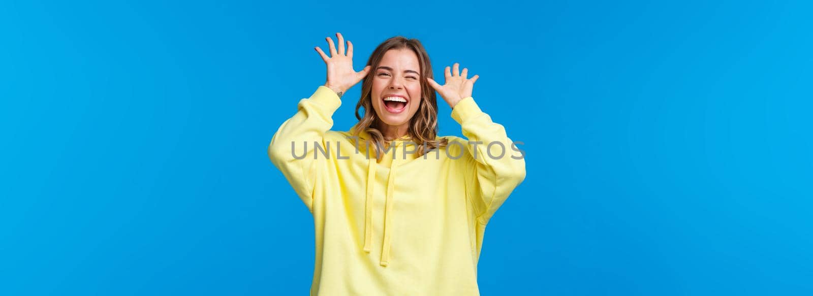 Try catch me. Alluring carefree european female with blond hair having fun, showing funny faces, laughing and smiling joyful, playing, make childish grimaces mocking someone, blue background.