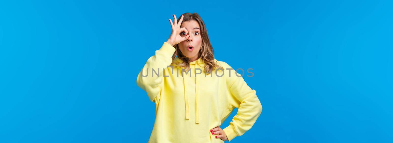 Very good, awesome. Impressed happy blond girl found something she was looking for, show okay gesture over eye and look through fingers, say wow astonished, stand blue background.